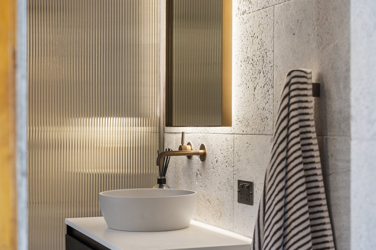 Bathroom vanity with a white vessel basin, brass wall-mounted tap, backlit mirror, textured stone tiles, and fluted glass panel