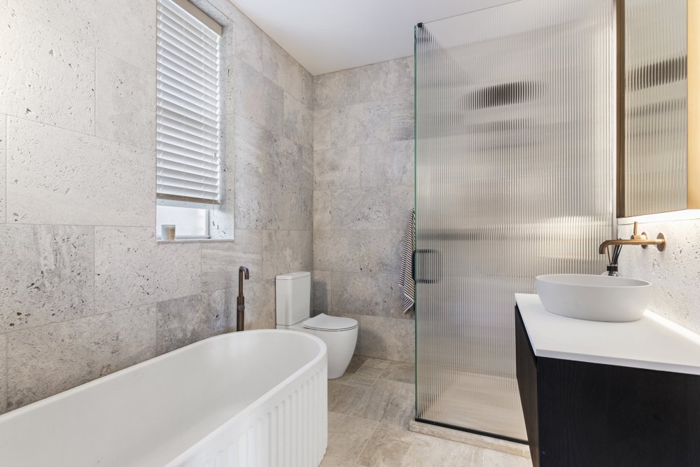 Modern bathroom with stone-tiled walls, freestanding bath, toilet, reeded glass shower screen, and vessel basin with brass tapware