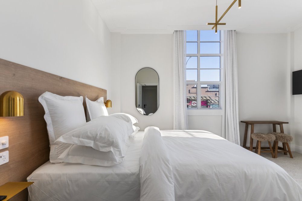 Bright hotel room with a king bed in white linen, timber headboard, gold wall sconces, oval mirror, and tall street-facing window