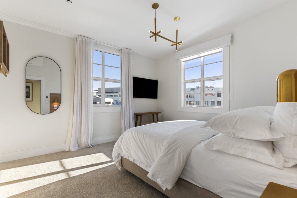 Bright hotel bedroom with a double bed, gold pendant light, wall-mounted TV, and large windows overlooking city buildings