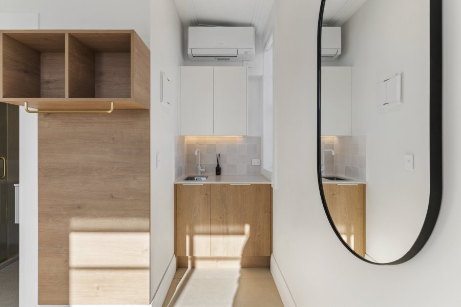 Compact kitchenette with oak timber cabinetry, white upper cabinets, tiled splashback, heat pump, and a round black-framed mirror