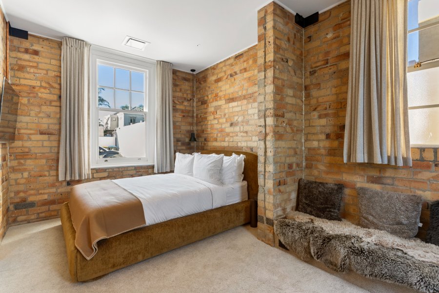 Bedroom with exposed brick walls, a double bed with white linen, and a cosy fur-draped seating nook beside a window