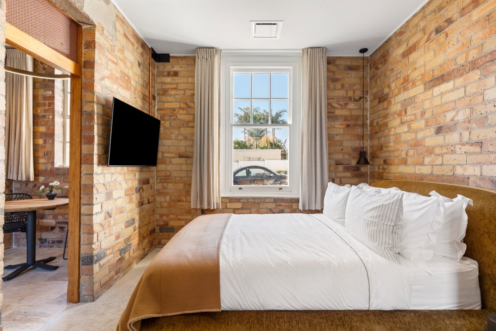Hotel room with exposed brick walls, double bed with white linen, wall-mounted TV, and a sash window with street views