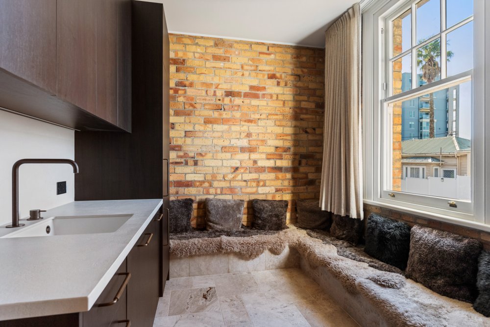 Interior with exposed brick wall, built-in window seat with fur cushions, stone benchtop, dark cabinetry, and city views through large sash windows