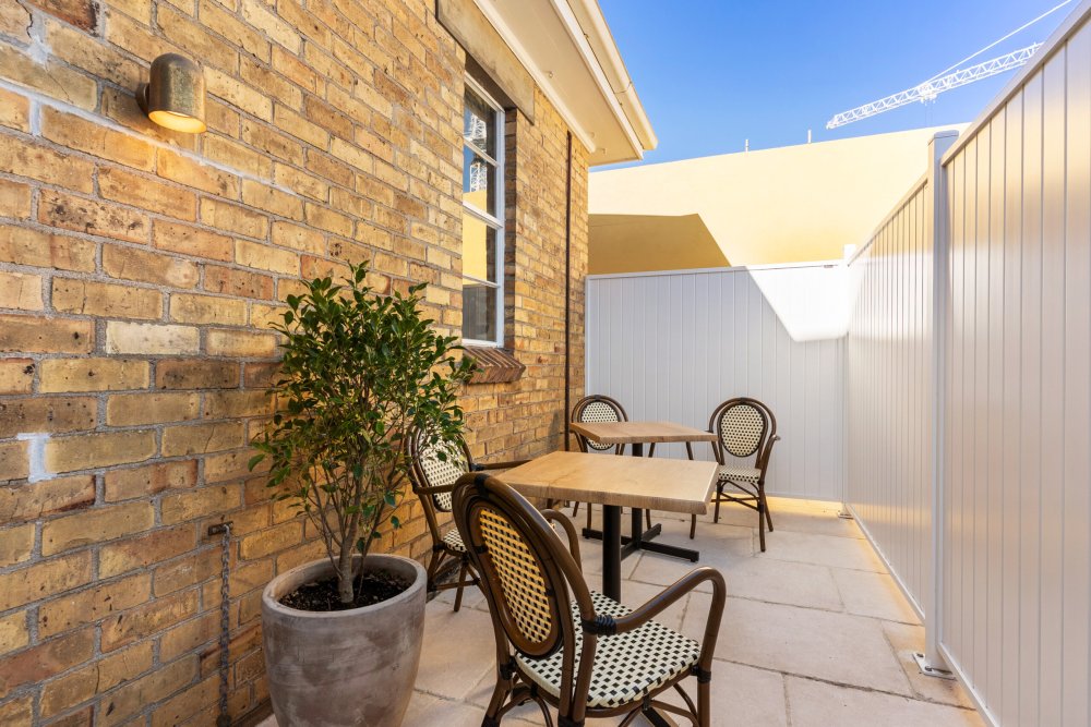 Outdoor courtyard seating area with bistro chairs and tables beside an exposed brick wall and potted plant under a clear blue sky