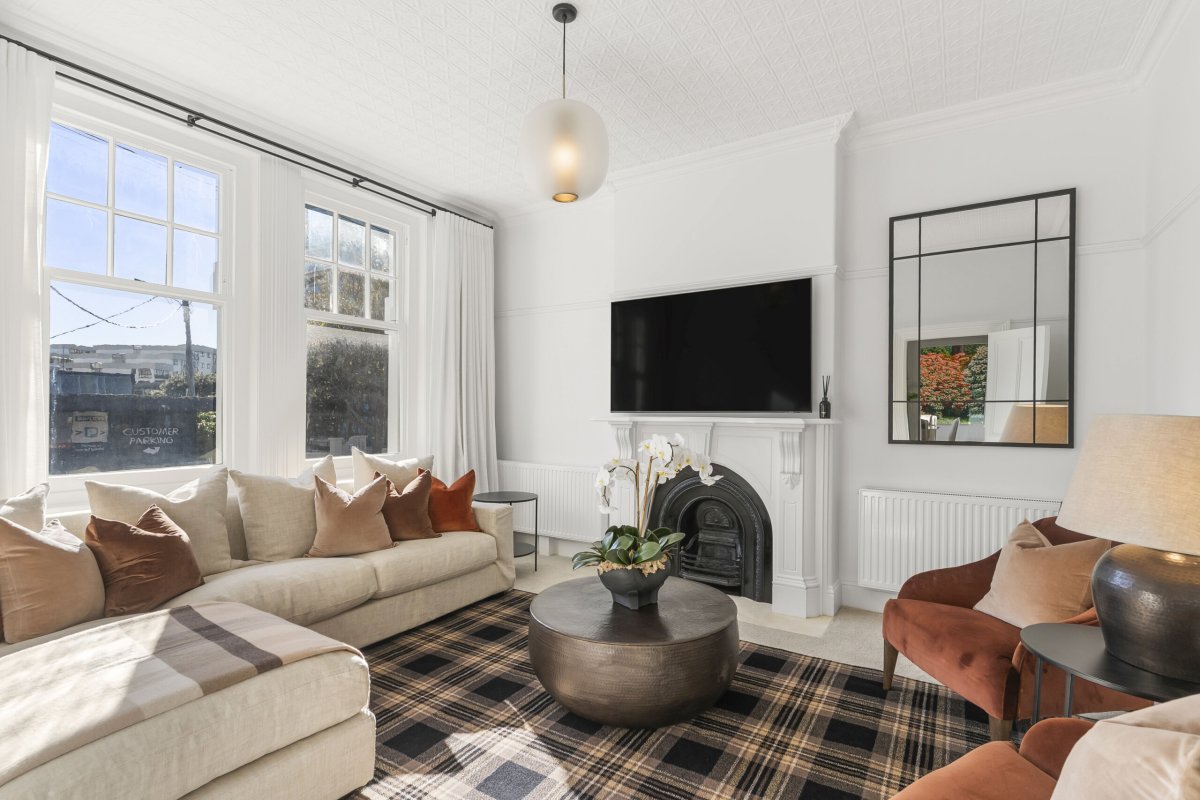Bright lounge with ornate fireplace, wall-mounted TV, cream sofa, rust armchair, and large sash windows